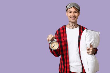 Mor arka planda pijamalı, yastıklı ve çalar saati olan dövmeli genç bir adam.