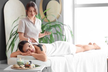 Masaj terapisti spa salonunda genç bir kadının sırtına çamur sürüyor.