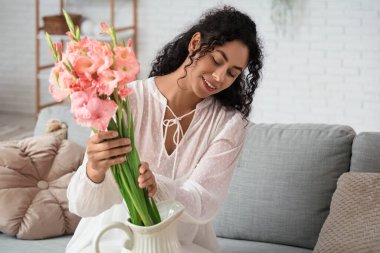 Güzel, genç, mutlu, Afro-Amerikan bir kadın evdeki vazoya pembe gladyatör çiçekleri koyuyor.