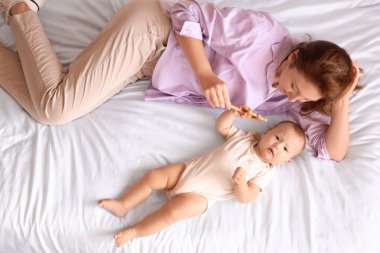 Güzel, genç bir anne ve küçük sevimli bir bebek yatak odasında uzanırken çıngırakla oynuyorlar.