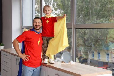 Mutlu bir baba ve küçük oğlu evdeki mutfağın penceresinin yanında süper kahraman gibi giyinmiş.