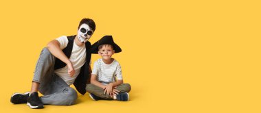 Baba ve küçük oğlu Cadılar Bayramı için giyinmiş korsanlar gibi sarı arka planda oturuyorlar.