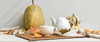 Çaydanlık, bir fincan sıcak içecek, balkabağı ve sonbahar yaprakları ışık duvarının yanındaki masada.