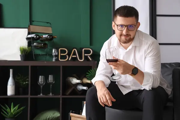 stock image Young sommelier with glass of wine sitting on black armchair in living room