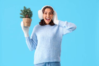 Mavi arka planda küçük bir Noel ağacı olan kalın elbiseli genç bir kadın.