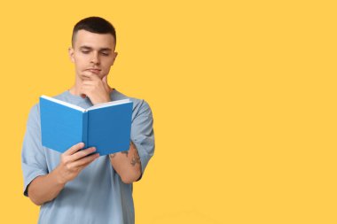 Düşünceli genç adam sarı arka planda kitap okuyor.