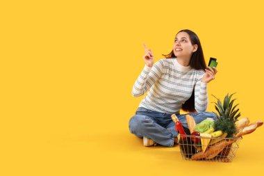 Alışveriş sepeti ve kredi kartları olan genç bir kadın sarı arka plandaki bir şeyi işaret ediyor.