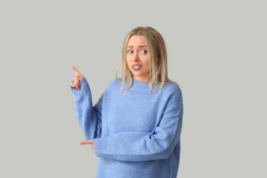 Young woman feeling awkward while pointing at something on grey background clipart