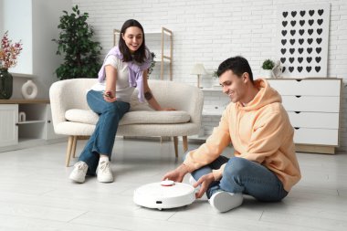 Mutlu genç çift oturma odasındaki modern robot elektrik süpürgesini uzaktan kontrol etmek için cep telefonu kullanıyor.