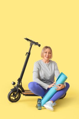 Güzel, olgun, sportif bir kadın. Sarı arka planda scooter ve yoga minderi var.