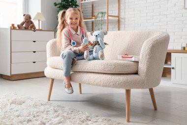 Evdeki kanepede ilk yardım çantası ve oyuncak ayısıyla oynayan tatlı küçük bir kız.