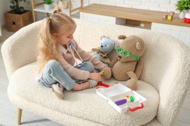 Evdeki kanepede ilk yardım çantası ve oyuncak ayıları olan sevimli küçük bir kız.