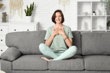 Evdeki kanepede oturmuş meditasyon yapan genç bir kadın.