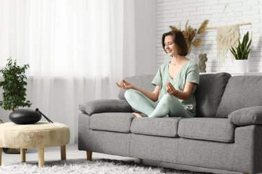 Evdeki kanepede oturmuş meditasyon yapan genç bir kadın.