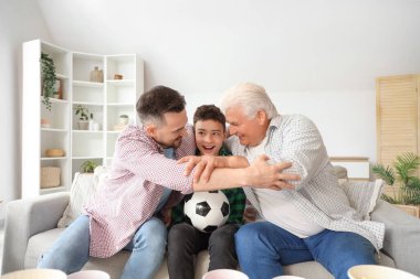 Genç bir çocuk, babası ve büyükbabasıyla evde maç izlerken sarılıyor.
