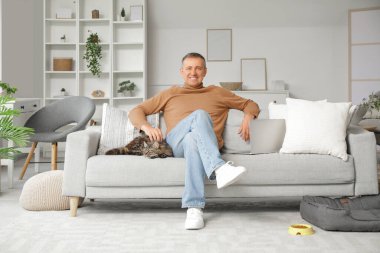 Evde kanepede oturan kedisi olan olgun bir adam.