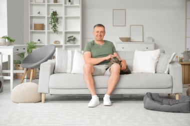 Olgun bir adam evdeki kanepede kediyi okşuyor.