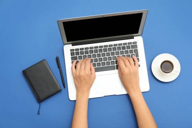 Dizüstü bilgisayarı, defteri ve mavi arka planda bir fincan kahvesi olan kadın elleri.