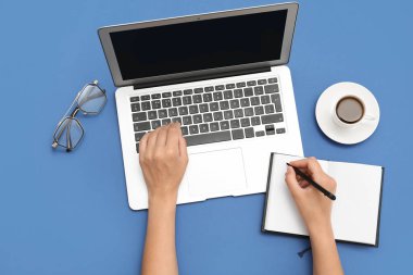 Dizüstü bilgisayarı, defteri ve mavi arka planda bir fincan kahvesi olan kadın elleri.