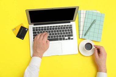 Dizüstü bilgisayarı, kredi kartları ve sarı arka planda bir fincan kahve olan erkek eller.