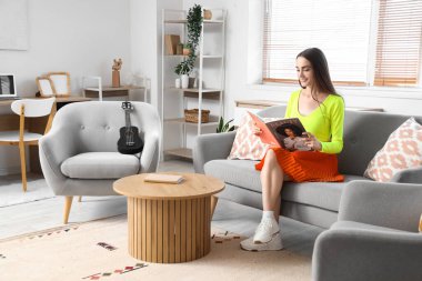 Genç ve güzel bir kadın evdeki kanepede dergi okuyor.