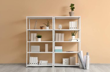 Ofis klasörleri ve oturma odasındaki bej duvarın yanında çiçekleri olan raf ünitesi.