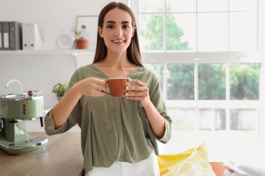 Güzel, genç, mutlu bir kadın, elinde bir fincan kahveyle mutfakta mola veriyor.