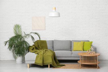 Asılı lamba, ekoseli yumuşak kanepe, çiçek, resim ve sehpa beyaz tuğla duvarın yanında.