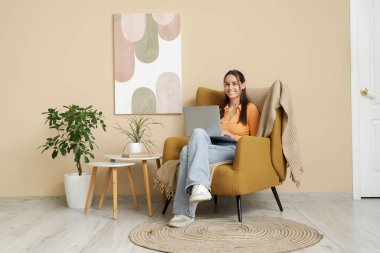 Genç bir kadın dizüstü bilgisayar kullanıyor ve oturma odasının içindeki bej duvarın yanındaki koltukta oturuyor.