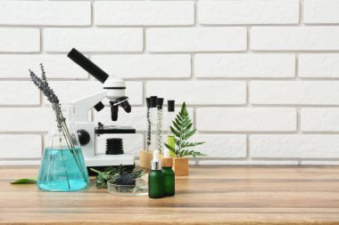 Beyaz tuğla duvarın yanındaki ahşap masada doğal kozmetik malzemelerinin hazırlanması için gereken malzemelerle laboratuvar camları ve mikroskobu.