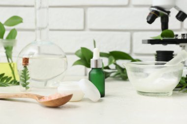 Tuğla duvarın yanındaki beyaz masada doğal kozmetik ürünlerini hazırlamak için gereken malzemelerle dolu bir laboratuvar cam ve mikroskobu.
