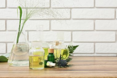 Beyaz tuğla duvarın yanındaki kahverengi ahşap masada doğal kozmetik ürünleri hazırlamak için malzemeler ve numunelerle dolu laboratuvar vitrinleri.