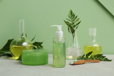 Yeşil duvarın yanındaki gri masada doğal kozmetik ürünlerini hazırlamak için malzemeler ve numunelerle dolu laboratuvar vitrinleri.