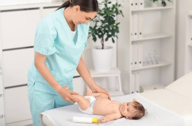 Küçük şirin bebek, klinikteki kanepede bir şişe kozmetik ürünüyle mutlu profesyonel terapistten bacak masajı yaptırıyor.