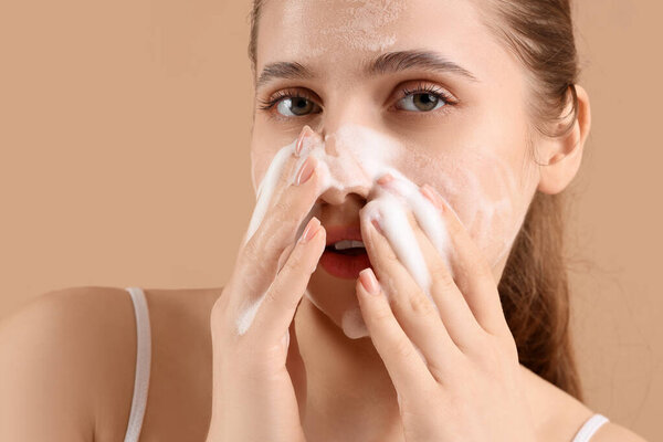 Beautiful young woman applying cosmetic foam on her face against brown background, closeup