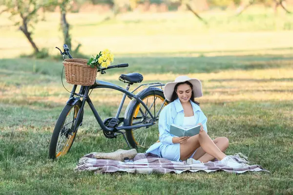 Bisikleti ve çiçekleri olan güzel, genç bir kadın parkta, açık havada ekose kitap okuyor.