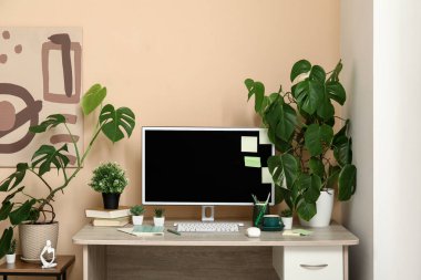 Evdeki bej duvarın yanında bilgisayar, kitap ve farklı ev bitkileriyle şık bir iş yeri.