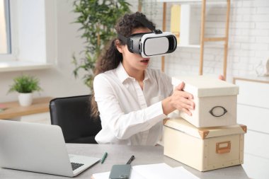 VR gözlüklü, ofisinde kutuları olan genç bir Afro-Amerikalı iş kadını.