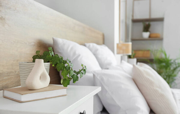 Vase with green plant and book on bedside table near bed in light bedroom, closeup
