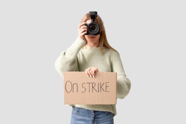 Gri arka planda, üzerinde metin olan bir pankart ve fotoğraf makinesi olan kadın fotoğrafçıyı protesto ediyor.