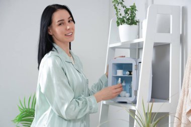 Genç bir kadın banyoda kozmetik buzdolabından serum alıyor.