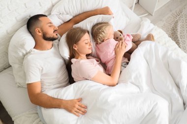 Ebeveynler ve oyuncak ayılı küçük kızları yatak odasında battaniyenin altında uyuyorlar.
