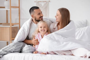 Mutlu ebeveynler ve yumuşak battaniyeli küçük kızları yatak odasında oturuyor.