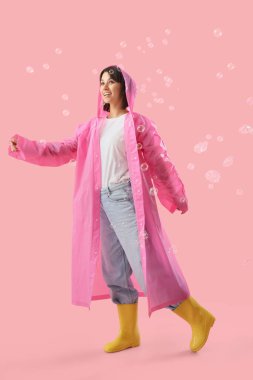 Yağmurluk ve pembe arka planda sabun köpüğü olan lastik çizmeli mutlu genç kadın.