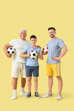 Babası ve dedesiyle birlikte sarı arka planda futbol topları tutan genç bir çocuk.