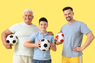 Babası ve dedesiyle birlikte sarı arka planda futbol topları tutan genç bir çocuk.