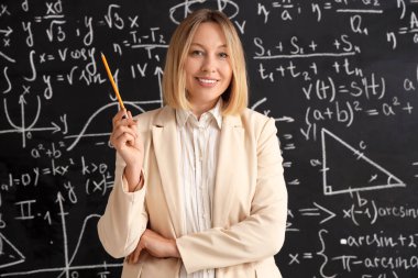 Sınıfta karatahtanın yanında kalem tutan olgun kadın matematik öğretmeni.
