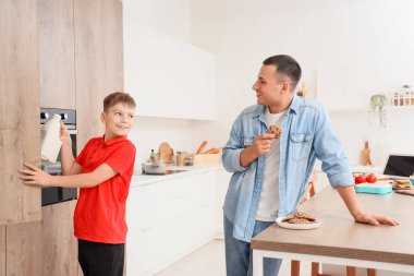 Küçük çocuk çekmeceden bir şişe süt alıyor ve babası da mutfakta kurabiye yiyor.