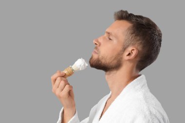 Sakallı genç adam gri arka planda fırçayla tıraş köpüğü sürüyor.