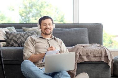Dizüstü bilgisayarı ve not defteri olan genç erkek öğretmen evde online ders veriyor.